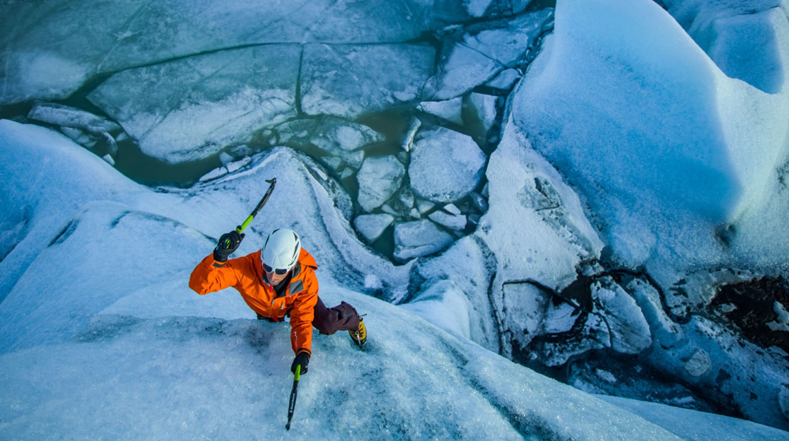 Klemin Premrl climbs up the side of a glacier in a bright orange jacket in Iceland while being photographed by Tim Kemple from above