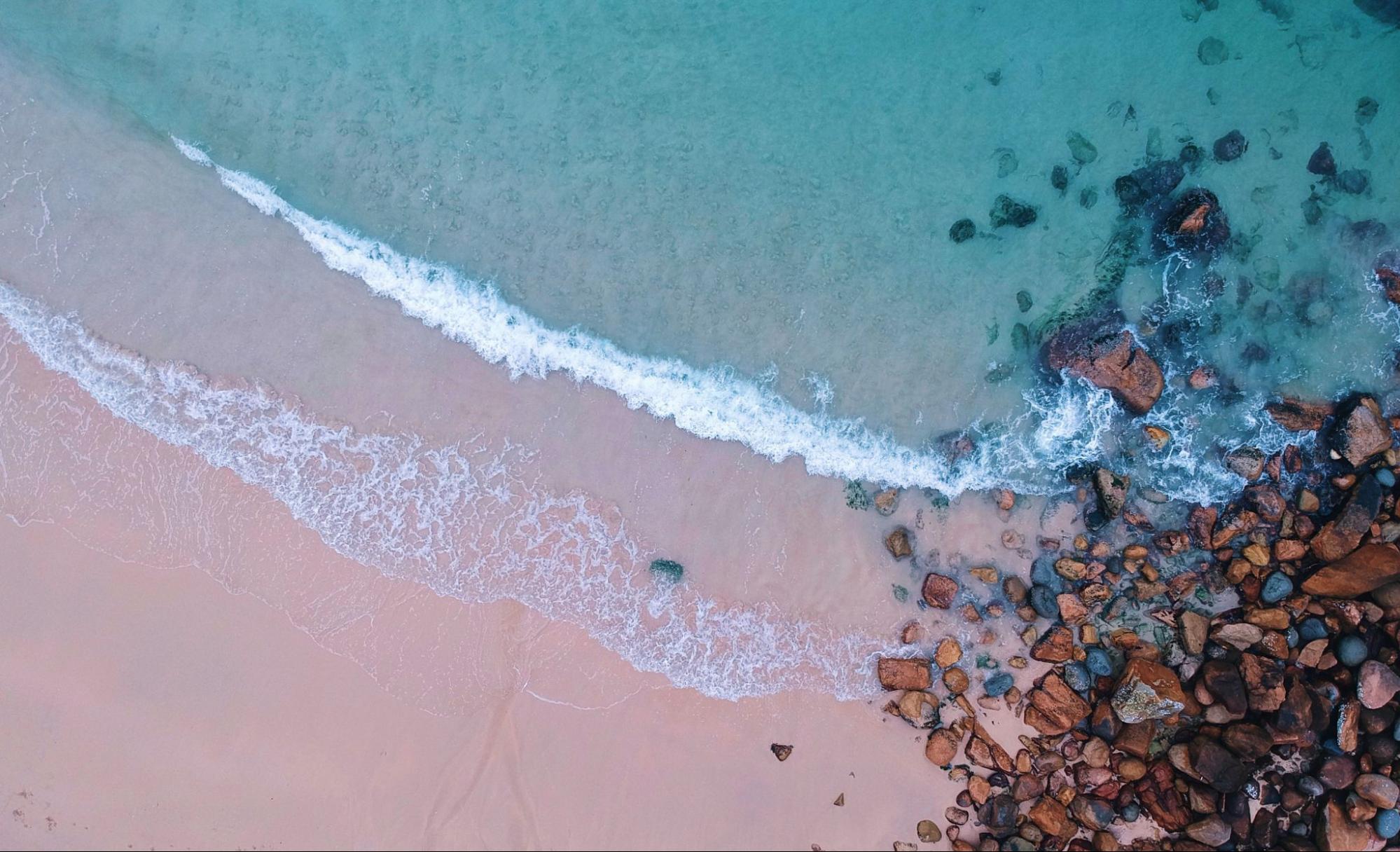 An overhead photo of a beach, with smooth sand transitioning into rocks on the shore and aquamarine waves rolling in. Photo by Taylor Simpson.