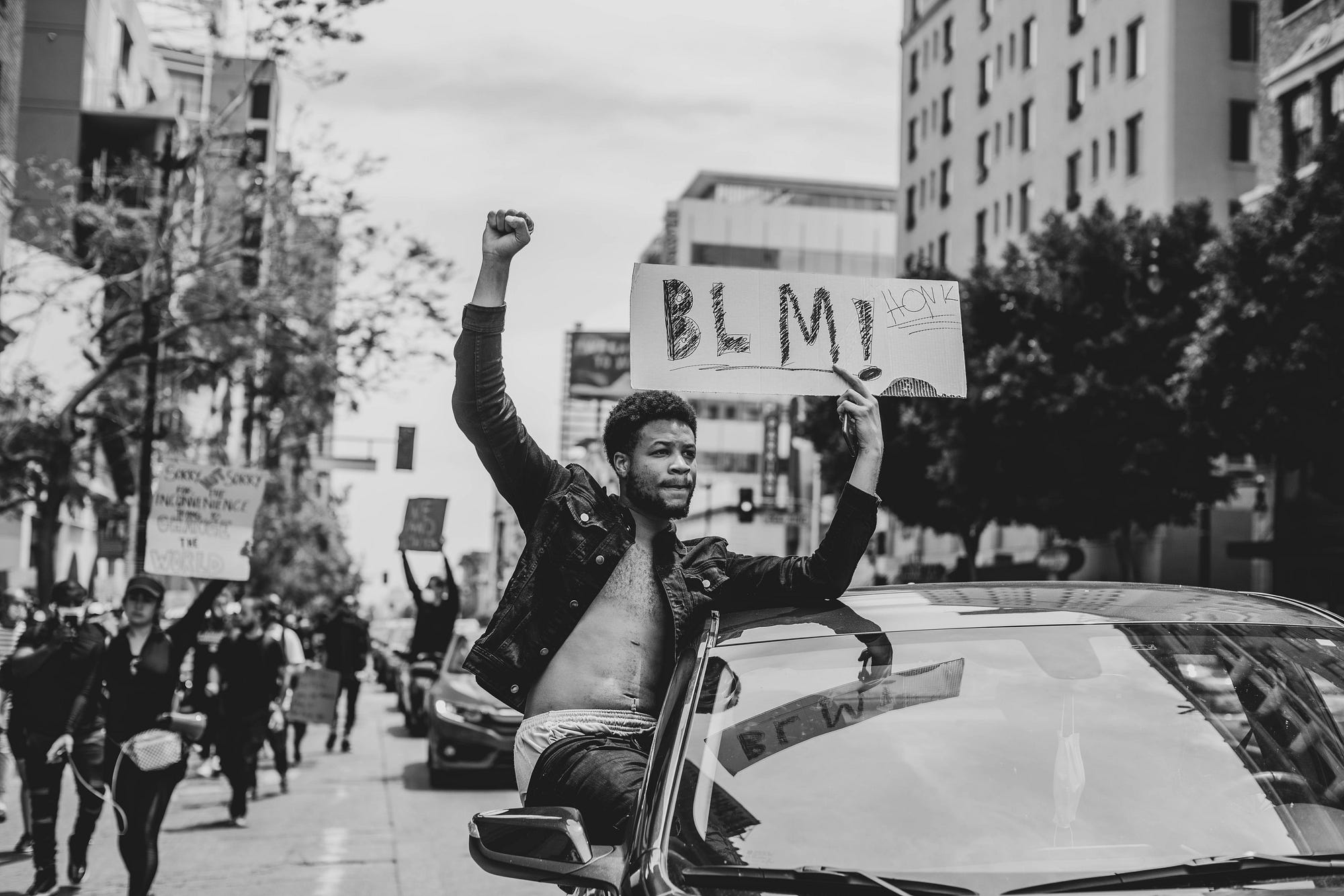 A Black man sitting in a car window holding up a fist and a Black Lives Matter sign.