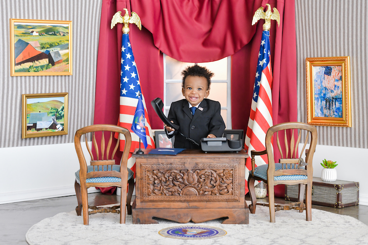 A baby dressed as the U.S. President standing behind a desk in a mock Oval Office.